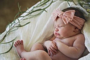 Sleeping baby girl in a pink bow headband lies on a white blanket beside green eucalyptus leaves in soft natural light.