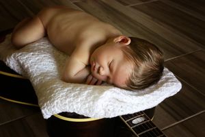 Newborn baby sleeps on a white crocheted blanket draped over an acoustic guitar on a rich wood floor.