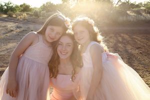 Three smiling girls in pink tulle dresses and flower crowns hug closely in golden sunset light in a desert field.