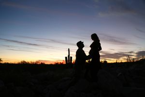 Silhouette of an expectant couple embracing against a colorful purple and orange desert sunset with a saguaro cactus nearby.