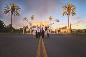 Family of six poses on a road lined with tall palm trees in front of a white temple under a pastel sunset sky.