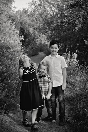 Black and white portrait of three smiling siblings standing together on a tree-lined outdoor path.