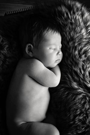 Black and white photo of a sleeping newborn baby curled up on a soft fur blanket.