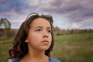 Young girl with wavy brown hair gazes upward in a grassy field beneath a dramatic cloudy sky.