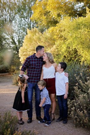 Smiling family of five posing outdoors among golden autumn trees as parents share a kiss.
