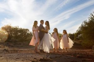 Bride and groom embrace at sunset in the desert while three young girls in pink dresses stand beside them.