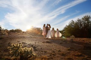 Bride and flower girls in pink dresses stand on a desert trail under a bright sunburst sky.