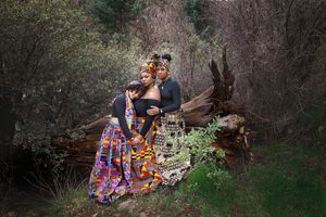 Three women in colorful African print outfits and headwraps pose together in a wooded forest setting.