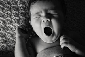 Black and white close-up of a newborn baby yawning widely with tiny fists raised.