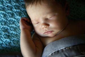 Sleeping newborn baby with one hand raised rests peacefully on a teal knit blanket in soft light.