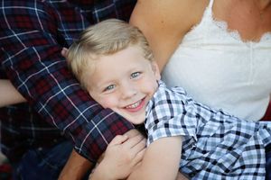A smiling blond boy in a plaid shirt hugs a parent's arm during a warm family portrait session.