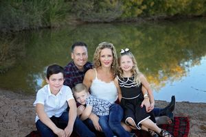 Family of five sits together on a red blanket beside a reflective pond with autumn trees, smiling for a portrait.
