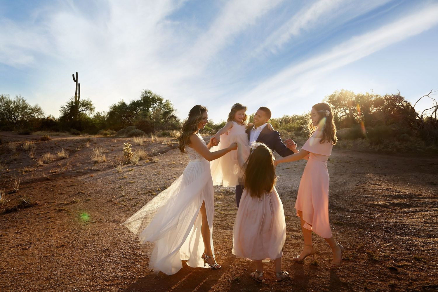 Bridal party in flowing white and pink dresses gathered in a joyful circle in the desert at golden hour.