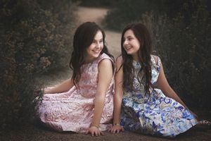 Two girls in floral dresses sit on the ground laughing together in a soft, moody outdoor portrait with dark foliage.