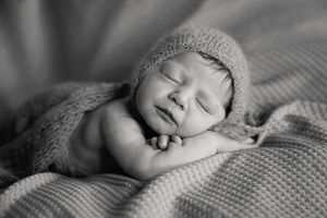 A black and white photo of a sleeping newborn in a knit bonnet resting its head on folded hands on a soft blanket.