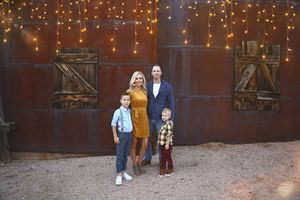 Family of four in coordinated outfits posing in front of a rustic rusted wall decorated with string lights.