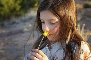 A girl with long hair gently smells a small yellow wildflower in the soft golden light of the desert.