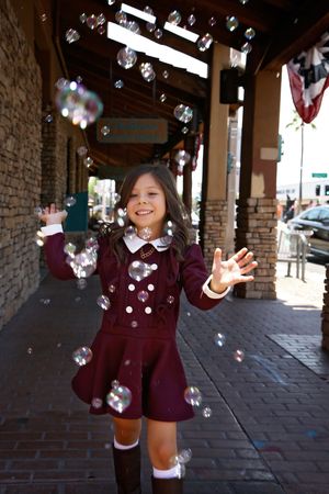 A smiling girl in a burgundy dress and boots plays with floating soap bubbles under a brick covered walkway.