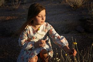 A girl in a floral dress crouches at sunset in the desert, touching small yellow wildflowers.
