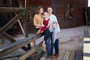 A family of four poses together on a rustic wooden rail, with parents smiling and their two young children in front of an old building.