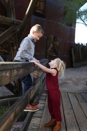 A young boy and a little blonde girl hold hands across a rustic wooden railing at an old western-style town.