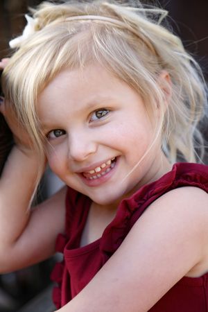 Close-up portrait of a smiling little blonde girl in a burgundy top with her hand resting on her head.