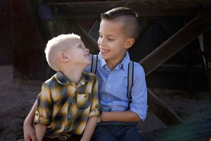 Two young brothers sit close together and smile at each other in front of a rustic wooden fence during a family photo session.
