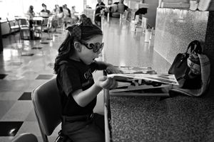 Black and white photo of a young girl in sunglasses and a bandana sitting at a table reading a magazine in a diner.