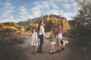 Mother and three daughters walk hand in hand on a desert trail with a small white dog and rugged mountains in the background.