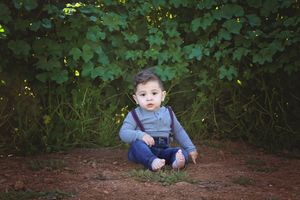Toddler boy in a gray shirt, suspenders and jeans sitting barefoot on the ground in front of leafy green vines.