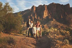 Family of five poses on a desert trail with rugged red mountain cliffs rising behind them at golden hour.