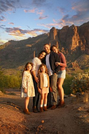Smiling family of five embraces on a desert path as sunset clouds glow above the rocky mountain backdrop.