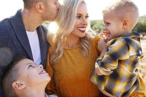 A smiling mother in a mustard sweater is kissed by her husband while holding her young son, with another boy looking up beside them.