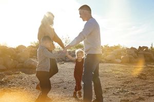 A family of four holds hands in a circle and plays in a sunlit desert landscape with rocks and golden sunflare.