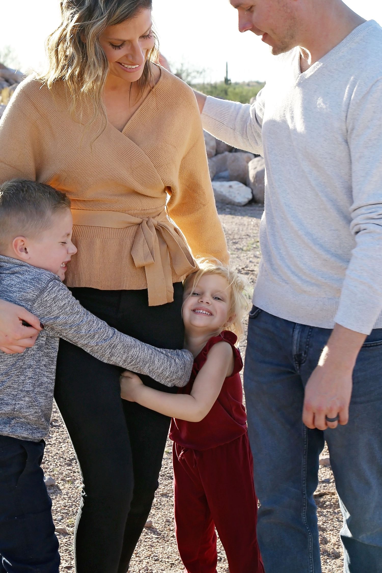 A mother in a tan wrap top smiles as her young son and daughter hug her, while the father stands close by outdoors.