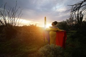 Pregnant woman in a flowing yellow and red gown standing in a desert landscape with a saguaro cactus at sunset.