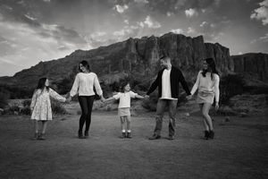 Black and white photo of a family of five holding hands in a line in the desert with rugged red mountains behind them.