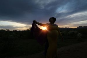 Silhouette of a pregnant woman in a long dress holding a flowing shawl against a dramatic sunset sky in the desert.