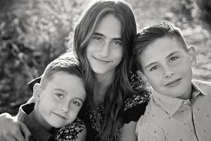 Black and white portrait of a teenage girl with two younger boys smiling closely together outdoors.
