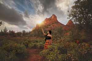 Woman in a colorful skirt reaches toward the sun among blooming desert shrubs with red rock formations in the background.