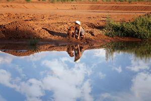 Child in a white sun hat crouches by a muddy red-dirt bank, reflected in a still pond mirroring puffy clouds.