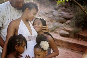 Tender family portrait of expectant parents embracing their three daughters among rocks and desert greenery.