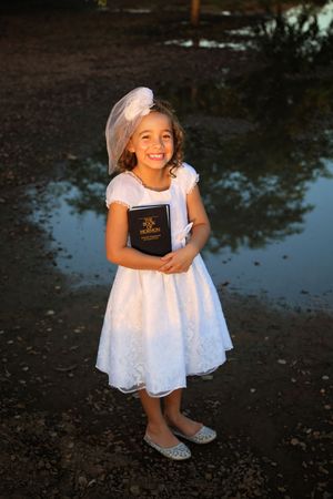 Smiling girl in a white lace dress and veil holds a Book of Mormon beside a reflective puddle at golden hour.