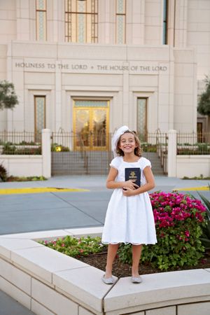 Smiling girl in a white dress and veil holds a Book of Mormon in front of the Holiness to the Lord temple entrance.
