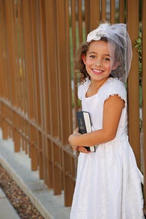Smiling girl in a white dress and floral veil holds scriptures while leaning against a tall wooden fence.