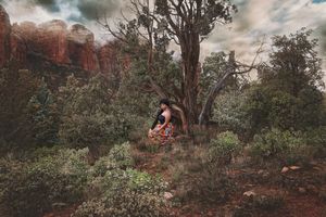 Woman in a colorful outfit sits beneath a gnarled tree among green shrubs with red rock cliffs in the background.