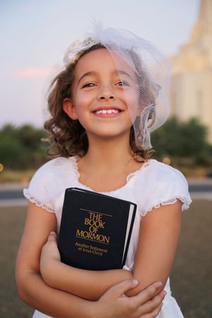 Smiling girl in a white dress and veil hugs a Book of Mormon at sunset with a temple behind her.