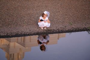 Young girl in a white dress and veil sits by a pool of water that reflects her and a temple.