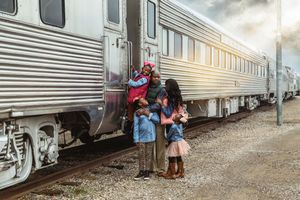 Family of four stands beside a silver passenger train on gravel tracks, with a father lifting a young girl under cloudy skies.