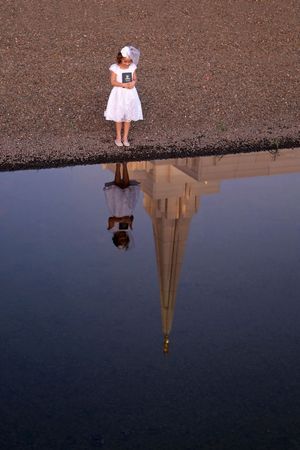 Girl in a white dress and veil holds a book beside still water reflecting her and a tall temple spire.